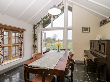 A dining room with a wooden table and chairs a vase with flowers a piano and large windows at Old Cary Cottage Henley near Langport