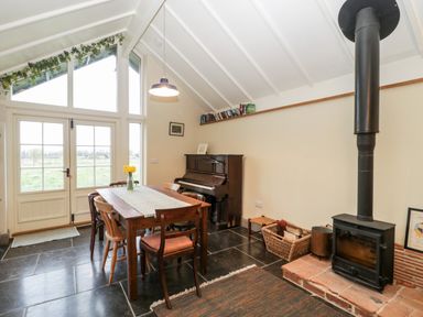 A dining area with a wooden table and chairs a piano a wood burning stove and large windows at Old Cary Cottage Henley near Langport