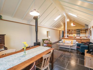 An open living space with dining table flowers wood stove armchairs sofa and kitchen shelves at Old Cary Cottage Henley near Langport