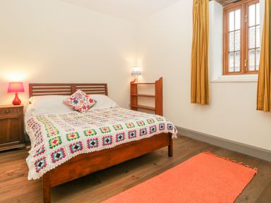 A bedroom with a wooden bed and colorful crochet blanket next to a wooden nightstand with a lamp and window with yellow curtains at Old Cary Cottage Henley near Langport