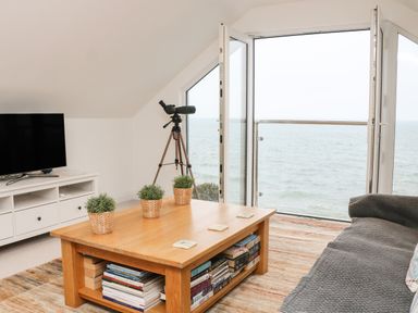 A living room with a gray couch wooden coffee table with plants and books a television on a white stand and a telescope by a window overlooking the sea at St Julien in Dawlish