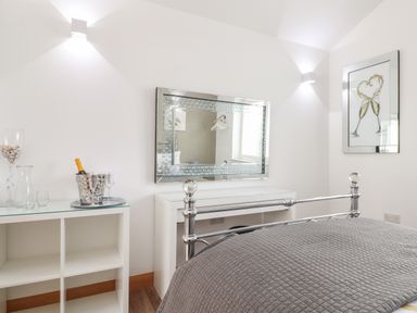 A bedroom with a metal bed frame a gray quilted bedspread a white dressing table with a large mirror and a shelf with a champagne bottle and glass at Chycoose Hicks Mill near Perranarworthal