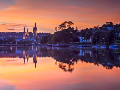 A river with boats and trees reflecting on the water with a cathedral and houses in the background at sunset