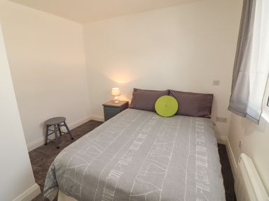 A bedroom with a bed and bedside table with a lamp a small stool and a window with curtains at Coolnaharragill Lower Annexe in Glenbeigh County Kerry