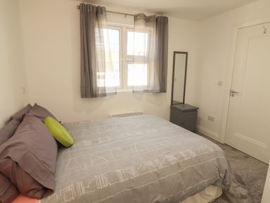 A bedroom with a bed gray pillows and a green round cushion a small side table with a mirror near a window with sheer gray curtains at Coolnaharragill Lower Annexe in Glenbeigh County Kerry