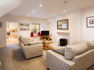A living room with two beige sofas a wooden coffee table a television on a stand a window with red cushions and a doorway to a kitchen at Fern Cottage in Ambleside
