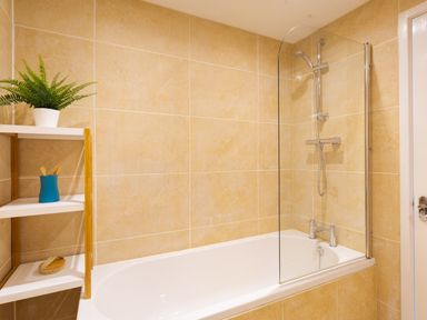 A bathroom with beige tiled walls a white bathtub a glass shower screen and a wooden shelf with a green plant and toiletries at Fern Cottage in Ambleside