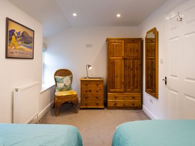 A bedroom with two twin beds a wooden wardrobe a chest of drawers a wicker chair with cushions a wall mirror and a framed picture at Fern Cottage in Ambleside
