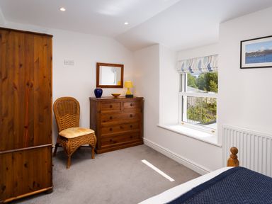 A bedroom with a wooden wardrobe chest of drawers a wicker chair and a window with a patterned blind at Fern Cottage in Ambleside