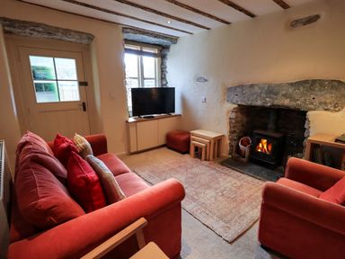 A living room with a red sofa red armchair a TV on a cabinet a stone fireplace with a wood burning stove and nested tables at Penny Red Cottage in Ambleside