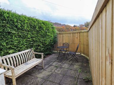 An outdoor patio area with a wooden bench a metal table and two chairs surrounded by a wooden fence and tall hedge at Penny Red Cottage in Ambleside