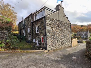 A stone exterior wall of a cottage at the corner of a paved road with trees and another house in the background at Penny Red Cottage in Ambleside
