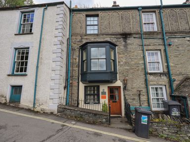 Exterior of stone and plaster houses with windows doors and trash bins on a sloped street at Heron Cottage in Ambleside