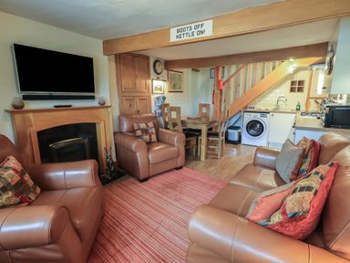 A living room with brown leather sofas and armchairs a wooden dining table under stairs an open plan kitchen with a washing machine and a wall mounted television at Heron Cottage in Ambleside