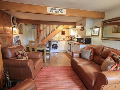 A living room with leather sofas a wooden dining table and chairs a kitchen with appliances under stairs at Heron Cottage in Ambleside