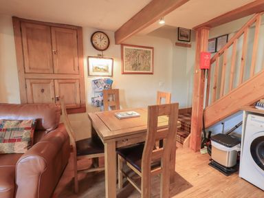 A dining area with a wooden table and four chairs next to a leather sofa and wooden staircase at Heron Cottage in Ambleside