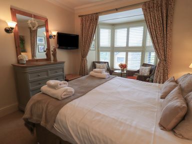 A bedroom with a bed folded towels a dresser a mirror wall lamps and a bay window with chairs at Heron Cottage in Ambleside