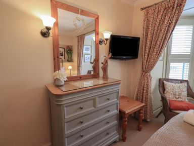 A bedroom corner with a chest of drawers mirror wall lights small table wicker chair and floral curtains at Heron Cottage in Ambleside