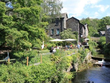 An outdoor seating area with umbrellas near a stone building by a stream surrounded by trees at The Miller in Ambleside