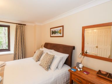A bedroom with a bed with pillows and a headboard next to a window with curtains and a wooden side table with a lamp a stuffed toy and a mirror on the wall at Waterfalls in Ambleside