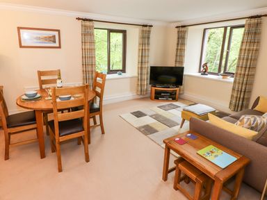 A living room with a wooden dining table and chairs a sofa a TV on a stand and two windows with curtains at Waterfalls Ambleside