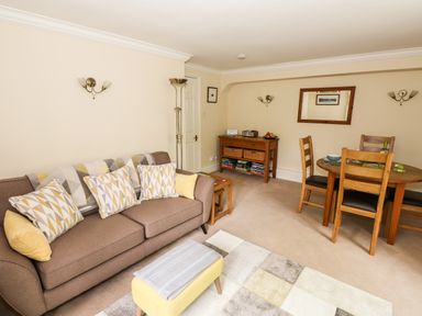 A living room with a brown sofa with cushions a small yellow ottoman a wooden side table and a dining table with chairs at Waterfalls in Ambleside