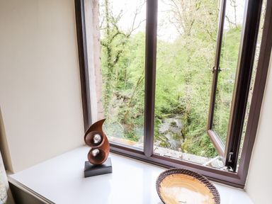A window overlooking a forest and stream with a wooden sculpture and a decorative bowl on the windowsill at Waterfalls in Ambleside