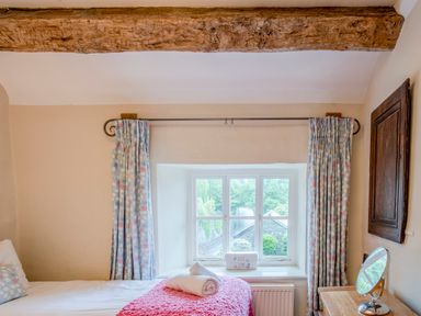 A bedroom window with floral curtains a wooden beam above and a bed with a pink throw at Knotts Cottage in Troutbeck