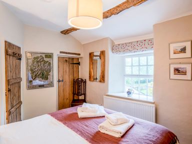 A bedroom with a bed, wooden doors, a window with a floral valance, a wooden chair, a mirror, and framed pictures at Knotts Cottage in Troutbeck