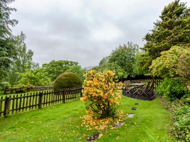 A garden with green grass a bush with orange flowers a wooden fence and outdoor table and chairs at Knotts Cottage in Troutbeck