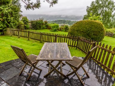 A wooden table with three chairs on a wet stone patio overlooking a garden with a wooden fence and trees at Knotts Cottage in Troutbeck