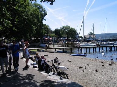 A waterfront with people walking and sitting near a beach with boats docked at piers and birds on the shore at Todd Crag in Ambleside