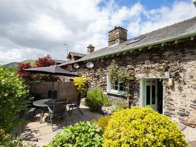 An outdoor patio area with a round table and chairs under an umbrella beside a stone cottage with greenery and a garden fence at Lowfold Cottage in Ambleside