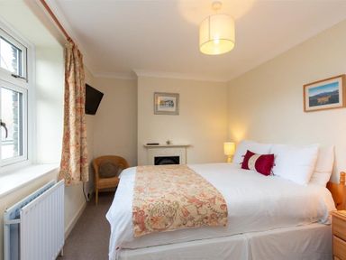 A bedroom with a double bed bedside table lamp wicker chair wall-mounted television and window at Sunset Cottage in Ambleside