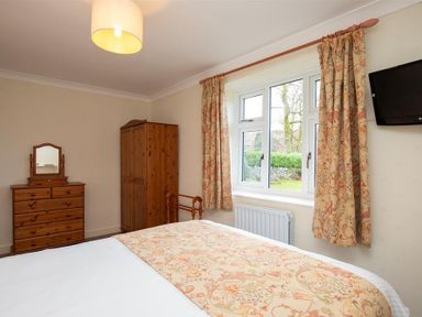A bedroom with wooden dresser wardrobe and bed with patterned bedspread and window with curtains at Sunset Cottage in Ambleside