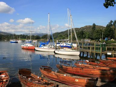 A lake with wooden rowboats and sailboats docked near a wooden pier with hills and trees in the background at Sunset Cottage in Ambleside