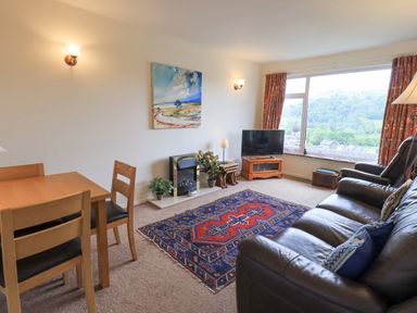 A living room with a wooden dining table and chairs a leather sofa and armchair a colorful rug a tv on a wooden stand and large window at Fellcroft in Troutbeck