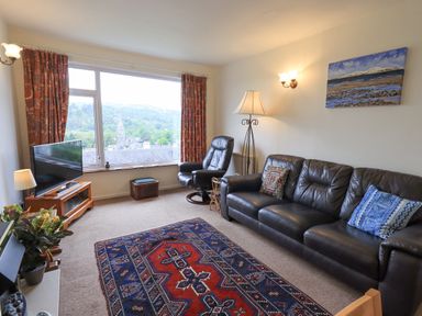 A living room with a leather sofa two armchairs a patterned rug a television and a large window showing a village view at Fellcroft in Troutbeck