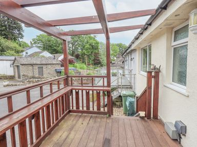 A wooden deck with railing and pergola outside a house with windows and a green trash bin at Fellcroft in Troutbeck