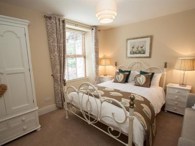 A bedroom with a white metal bed frame two bedside tables with lamps a wardrobe and window with patterned curtains at Acorn Cottage Ambleside