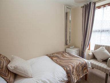 A bedroom with a single bed brown patterned blanket a white side table with a bowl a tall mirror striped curtains and an armchair with cushions at Acorn Cottage Ambleside