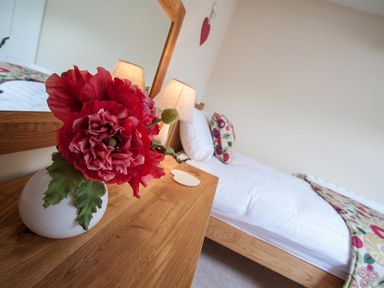 A bedroom with a wooden dresser holding a white vase with red flowers and a lamp next to a bed with white bedding and floral cushions at Acorn Cottage in Ambleside