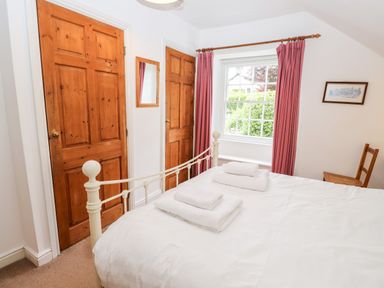 A bedroom with a white metal bed frame towels on the bed wooden doors a window with pink curtains and a wooden chair at Rock Cottage in Ambleside