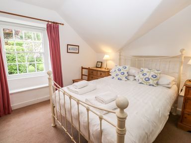 A bedroom with a bed with white linens and patterned pillows a wooden chest of drawers and a window with red curtains at Rock Cottage in Ambleside