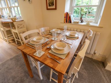 A dining table set for four with white chairs near a window with books and decorative items at Highfold Ambleside