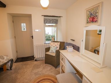 A room with a door a window a wicker chair with cushions a white dressing table with a mirror and a round rug on the carpeted floor at Highfold in Ambleside