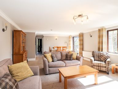 A living room with sofas, wooden coffee table, dining table with chairs, wooden cabinet, and windows with curtains at Ghyll View in Ambleside