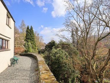 A gravel patio with green metal chairs and a stone wall next to a house with trees and shrubs in the background at Rawfell in Great Langdale