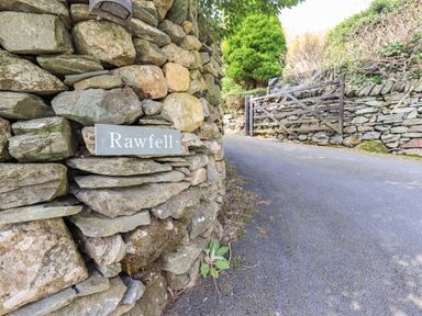 A stone wall with a Rawfell sign next to a paved road and a wooden gate at Rawfell Great Langdale
