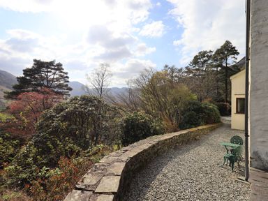 A curved stone wall beside a gravel pathway with two green metal chairs and a table next to a cream building surrounded by trees and bushes at Rawfell in Great Langdale
